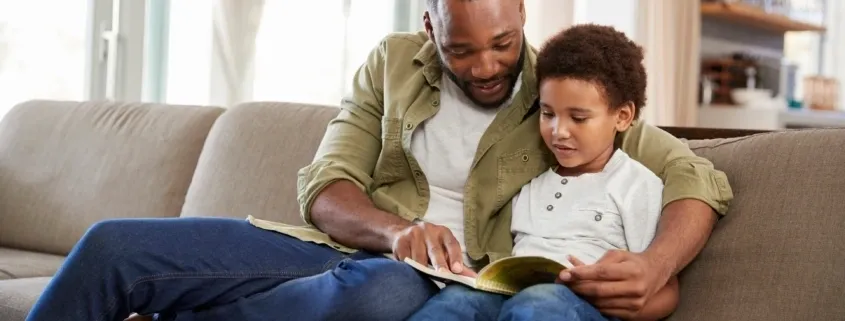 A man and child reading a book on the couch.