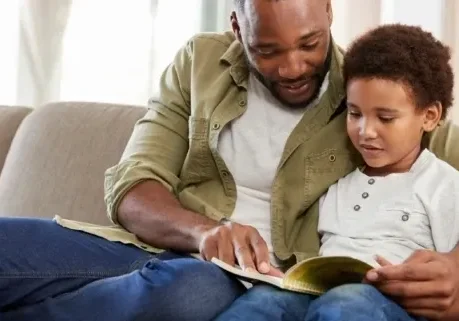 A man and child reading a book on the couch.